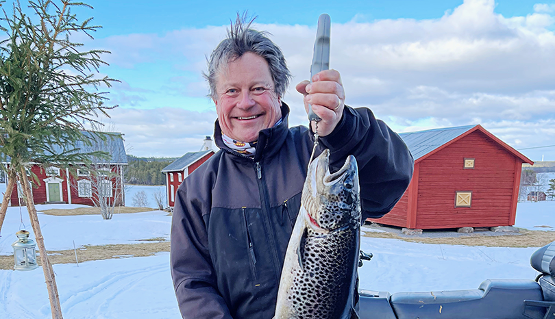 Man med stort  leende håller upp fisk. I bakgrunden två röda stugor.