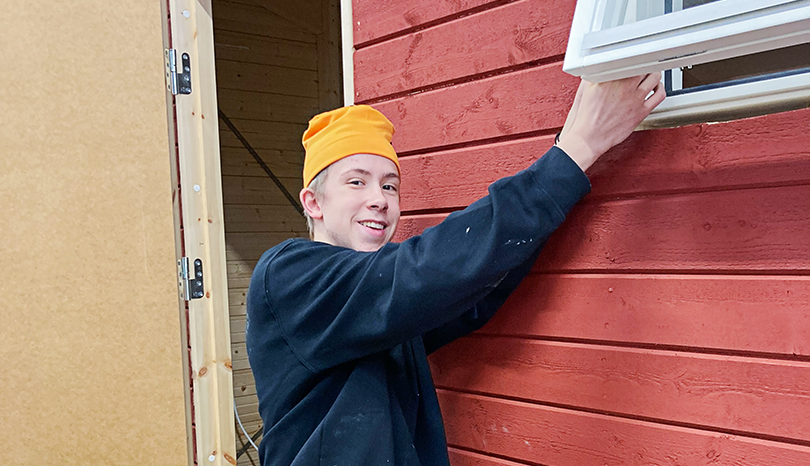 Kille i snickarkläder står vid rött hus.